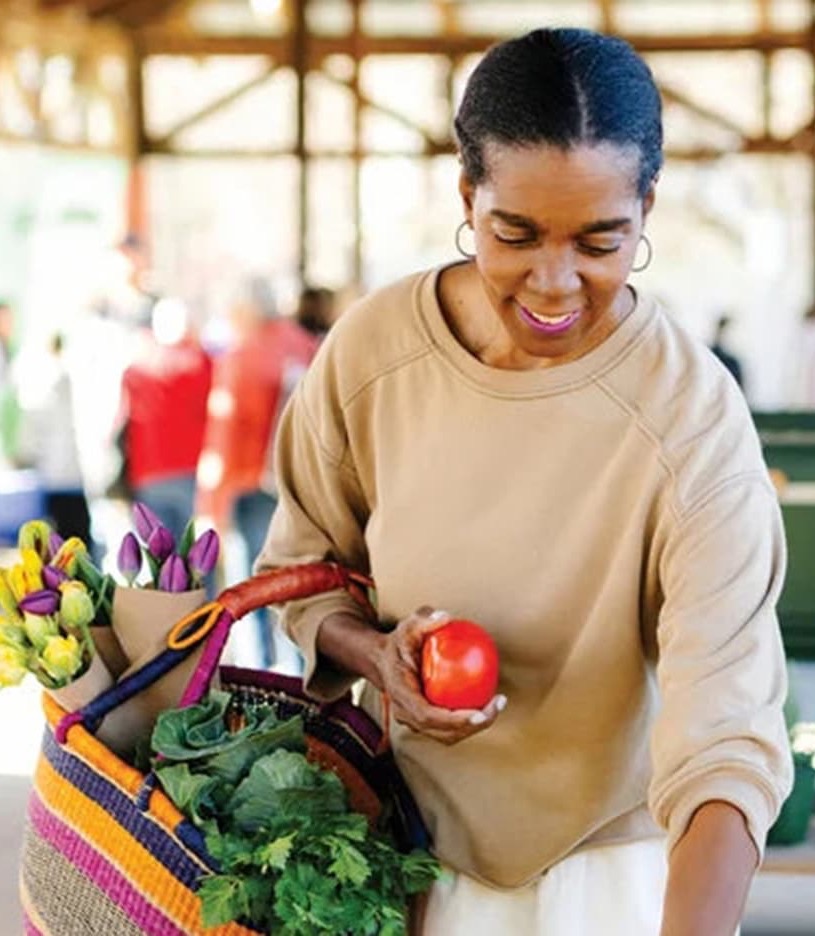 Spring Council shopping at a farmers market, holding a red tomato and a colorful tote bag filled with fresh greens and flowers.