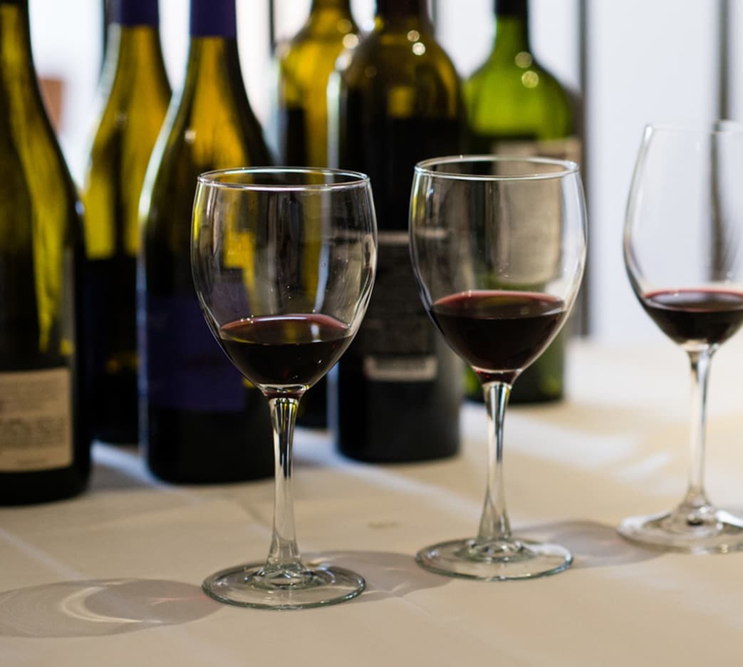Three wine glasses with red wine lined up on a table, with wine bottles blurred in the background.