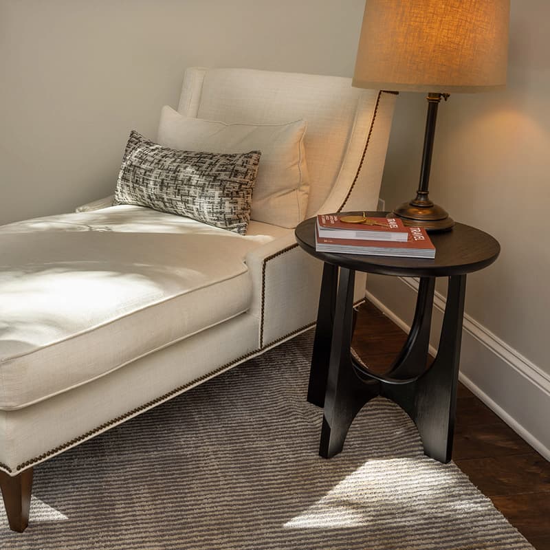 Upholstered lounge chair next to a small round table with a lamp and two books.