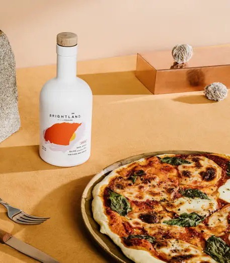A Margherita-style pizza with melted cheese and fresh basil sits on a plate beside a bottle of Brightland olive oil, set on a warm-toned tabletop with minimal, modern styling elements.