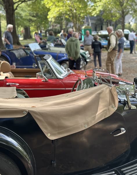Classic convertible cars parked with tan and red interiors, while people stand and chat in the background among trees on a sunny day. The scene suggests a car show or gathering in a park-like setting. Fearrington Village