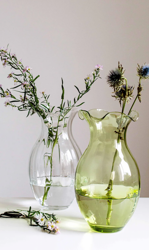 Two glass pitchers used as vases—one clear, one pale green—holding delicate wildflowers and thistle stems on a white surface against a neutral wall.