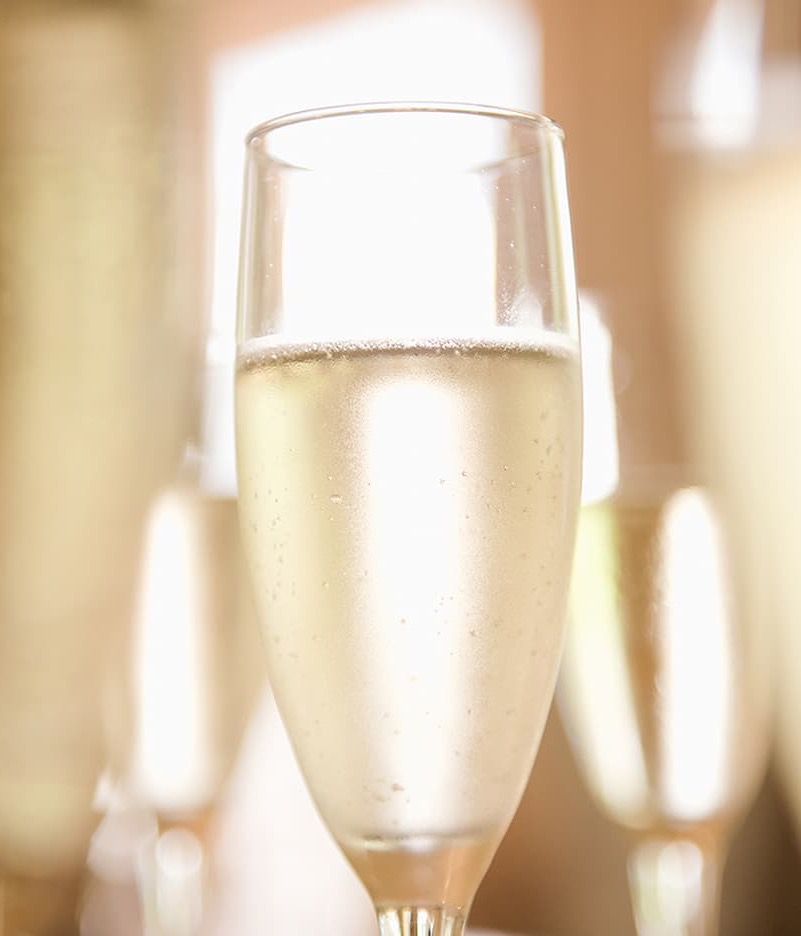 A close-up of a chilled glass of sparkling wine with tiny bubbles rising, softly lit with blurred glasses in the background.