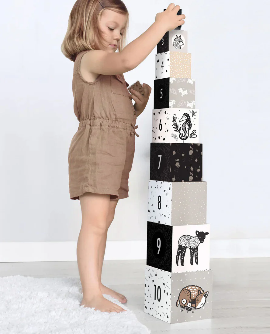 A young child stacks illustrated number blocks into a tall tower, each block featuring monochrome animals and patterns.