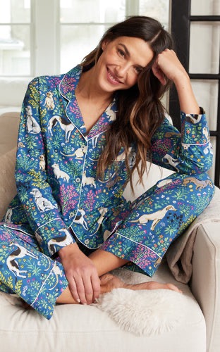 Smiling woman wearing a blue pajama set with playful animal and floral patterns, seated cross-legged in a cozy armchair indoors, resting her head on one hand in a relaxed pose.