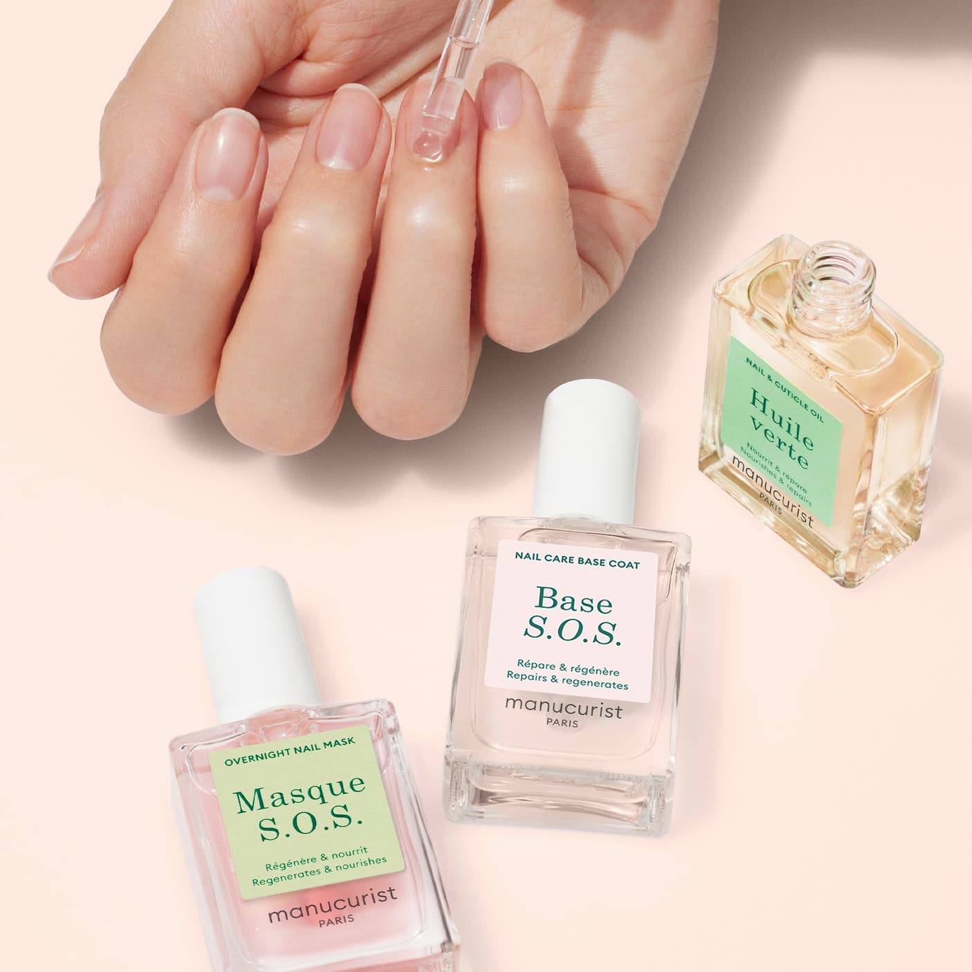 Hand applying a drop of nail oil with a glass dropper, surrounded by Manucurist nail care products including Base S.O.S., Masque S.O.S., and Huile Verte on a soft neutral background.