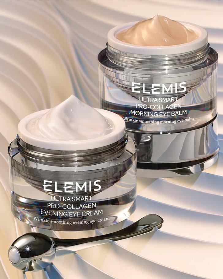 Two open ELEMIS Pro-Collagen eye cream jars with a metal applicator on a light, wavy background.