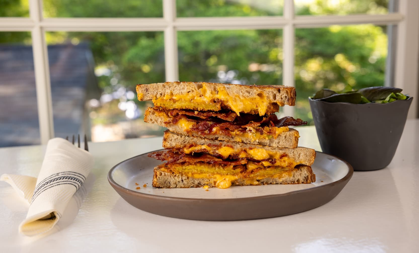 Stacked grilled bacon and cheddar sandwiches on toasted sourdough, filled with crispy bacon and melted sharp cheddar, served with a fresh green salad beside a sunlit window.
