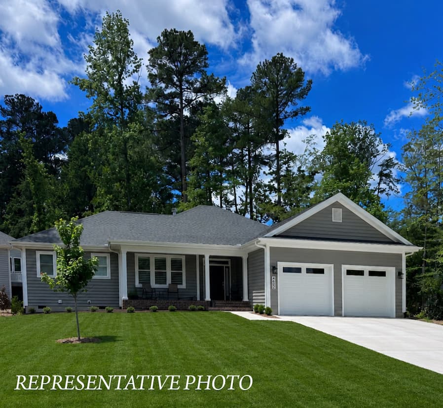 Gray single-story home with white trim and a two-car garage, set against a backdrop of tall pine trees and a bright blue sky with scattered clouds. A neatly manicured green lawn and young ornamental tree frame the front of the property, with “Representative Photo” displayed across the lower left corner.