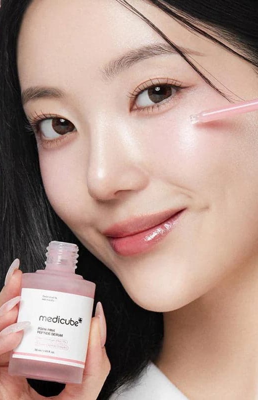 Woman applying Medicube PDRN Pink Peptide Serum with a dropper against a pink background. Fearrington Village.
