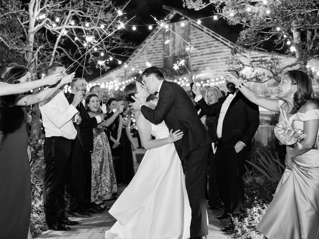 Black-and-white photo of a bride and groom kissing as guests hold sparklers around them outside a barn lit with string lights at night.