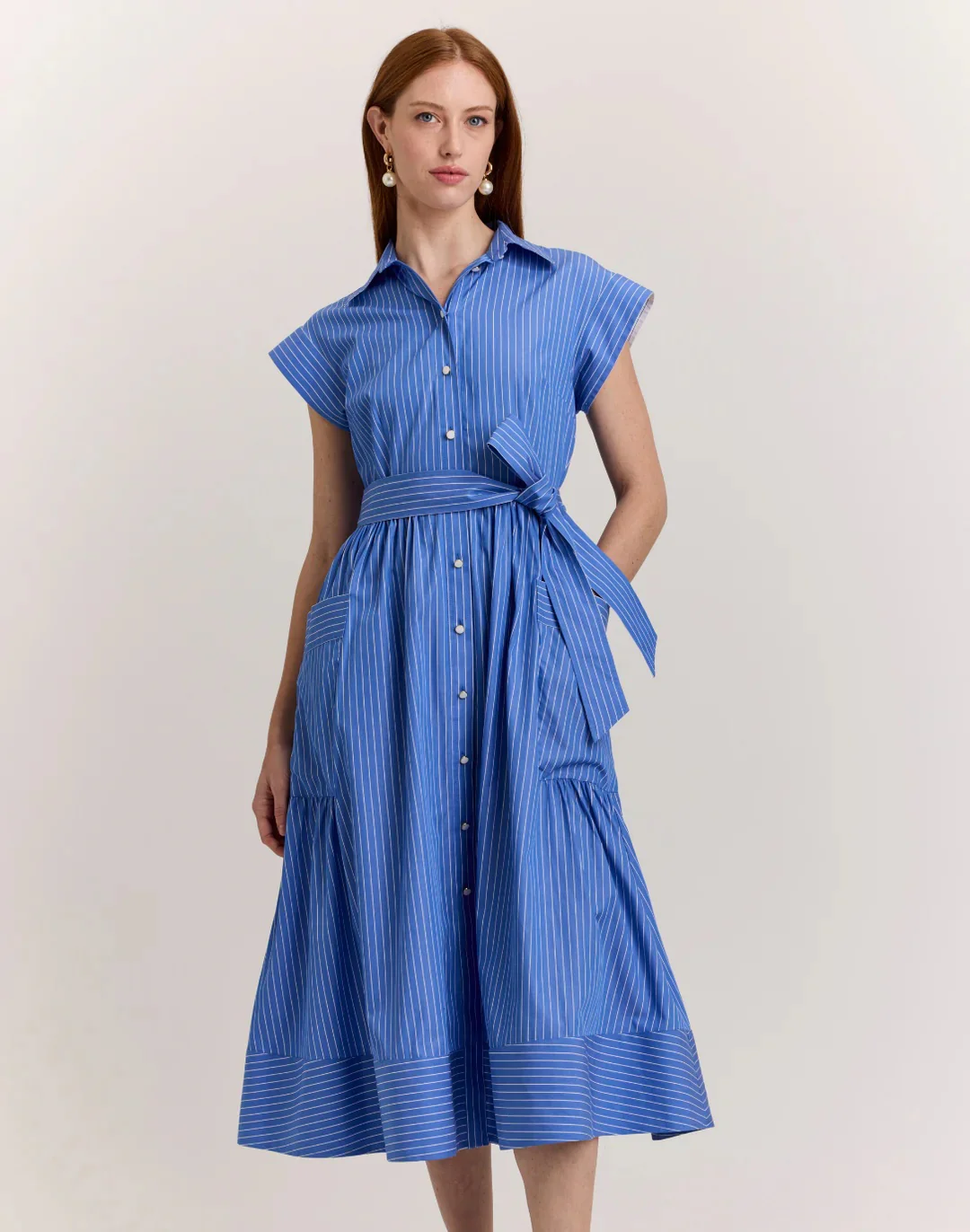 A woman models a blue, pinstriped shirt dress with cap sleeves, a button front, and a tied waist, styled with drop earrings against a neutral background.