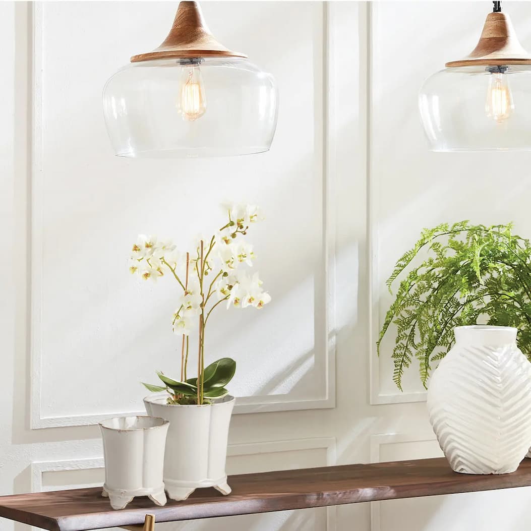 White orchids and a lush green fern add a fresh, modern touch to a console table styled with sculptural white ceramic planters and a textured vase beneath warm glass pendant lights.