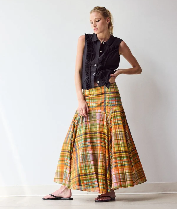 Woman wearing a sleeveless black button-front blouse with ruffle trim, paired with a full plaid maxi skirt in warm shades of gold, orange, pink, and green. The look is finished with black flip-flops against a neutral studio backdrop.