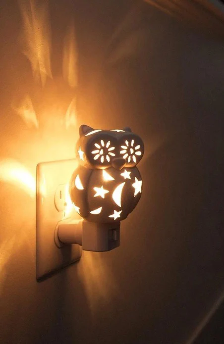 Owl-shaped plug-in nightlight glowing warmly in a wall outlet, casting star and flower patterns of light across the surrounding wall.