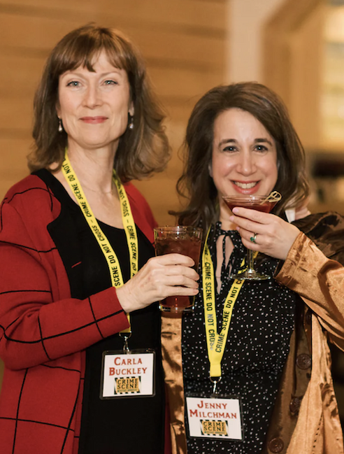 Two women standing indoors at an event, smiling and holding drinks, each wearing a name badge and a yellow lanyard, in a warm, softly lit setting.
