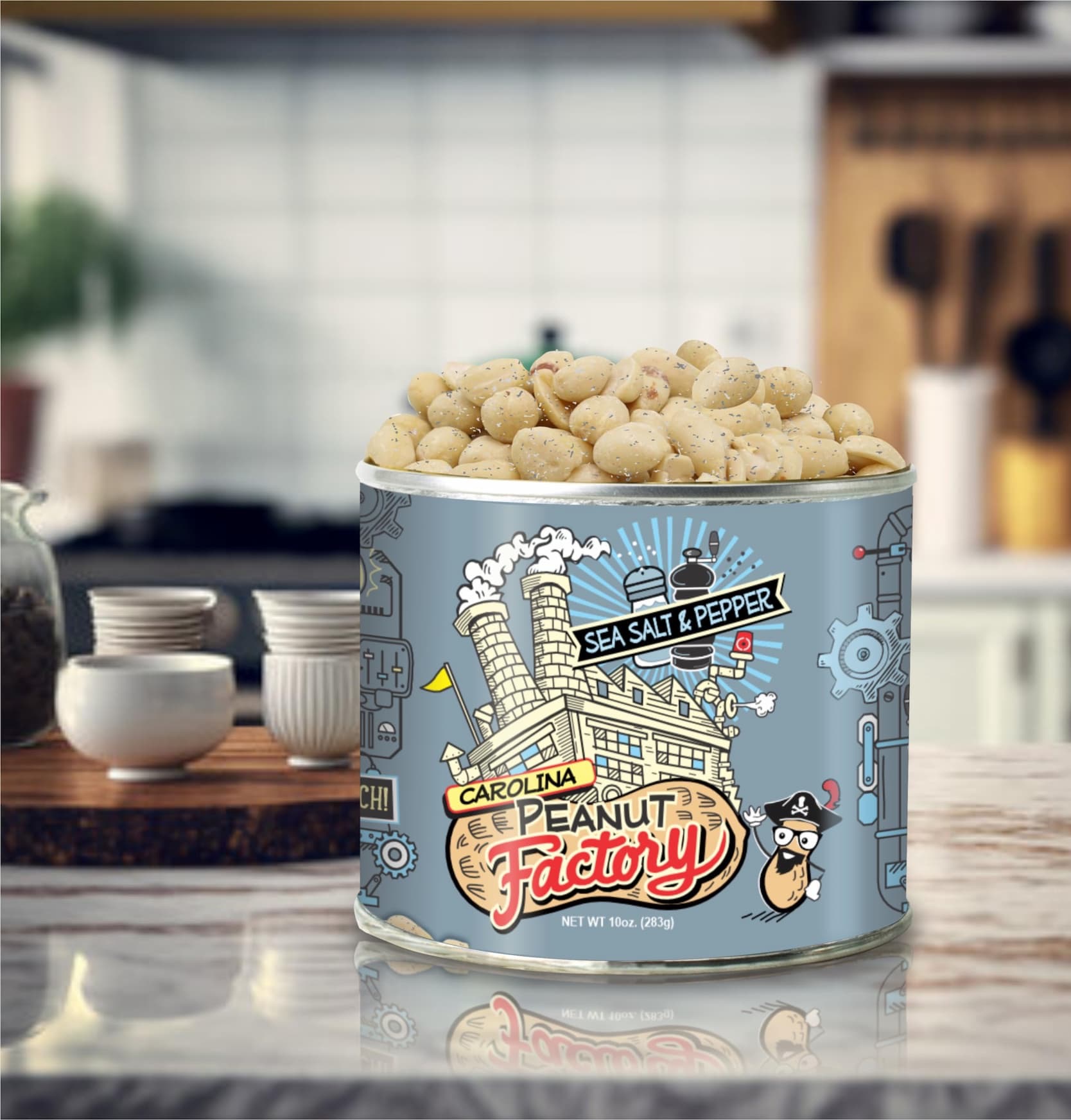 A tin of Carolina Peanut Factory Sea Salt & Pepper peanuts sits on a polished countertop in a bright kitchen. The vintage-inspired blue label features whimsical factory illustrations and a cartoon peanut mascot, while the tin is filled to the brim with lightly seasoned peanuts. Softly blurred kitchenware and neutral décor in the background create a warm, inviting setting.