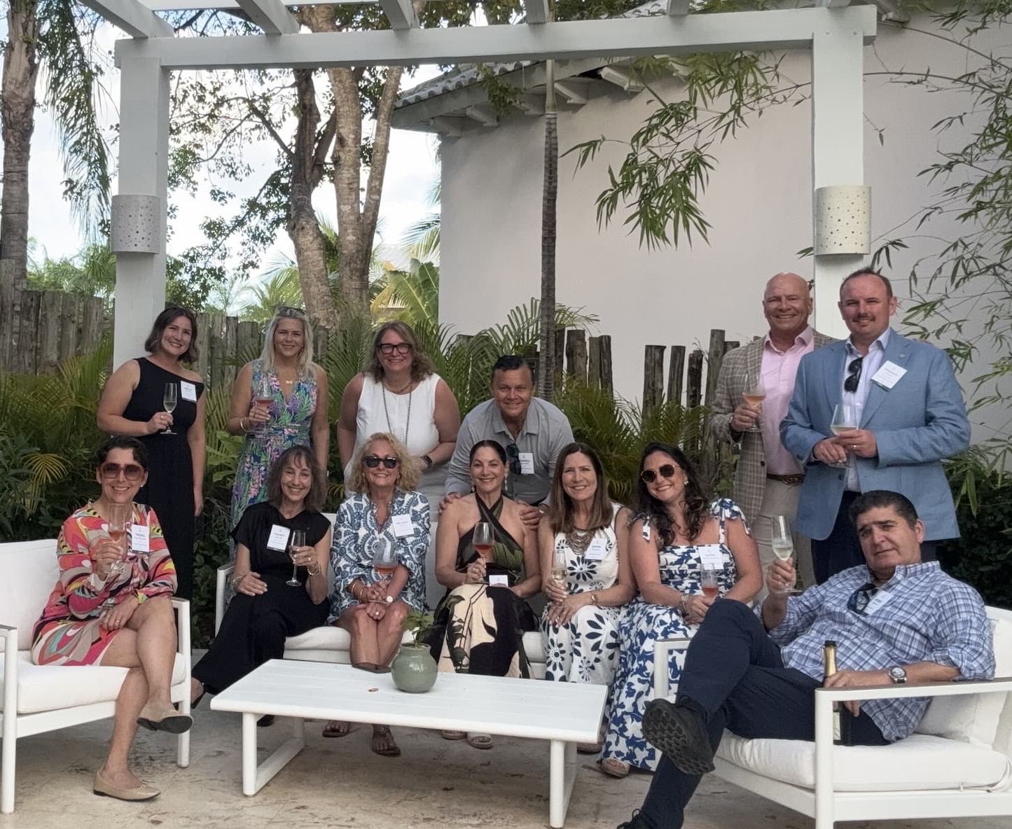 A group of well-dressed adults gathered outdoors under a white pergola, smiling and holding glasses of wine. They are seated and standing around modern white patio furniture, surrounded by lush greenery and tropical plants. The setting feels relaxed and social, with a mix of casual elegance and warm, late-afternoon light.