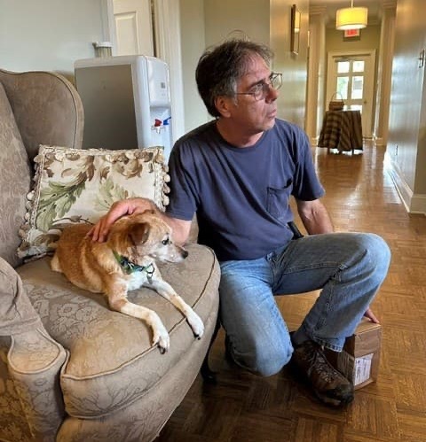 A man kneels beside a small tan dog resting on an upholstered armchair in a softly lit hallway. With one hand gently on the dog's back, he looks down the corridor while the dog relaxes comfortably on the cushion. Warm wood floors, neutral furnishings, and natural light create a calm, home-like atmosphere that highlights the close bond between the person and the dog.