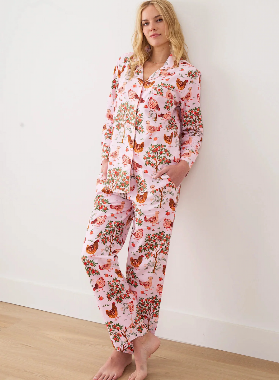 Person standing against a plain wall wearing a matching pink pajama set with a whimsical print of birds, trees, and fruit, hands in pockets.
