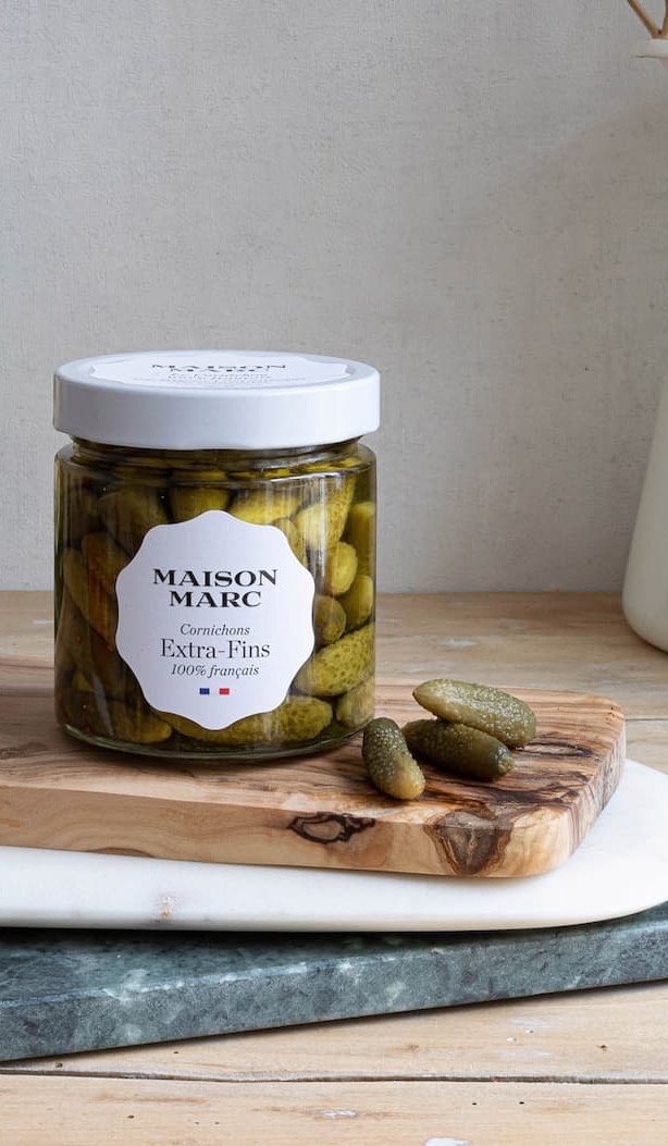 Glass jar of Maison Marc extra-fine French cornichons on a wooden board, with a few pickles beside it.