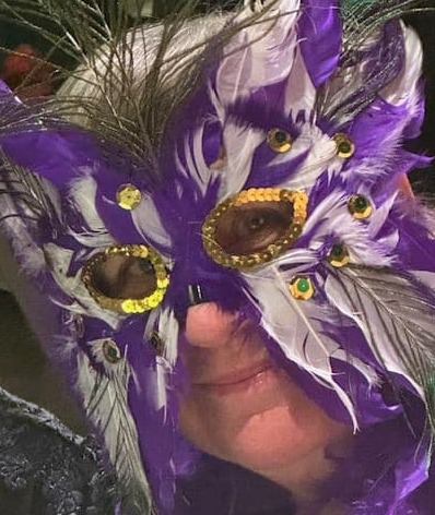 Person wearing an ornate purple and white feathered masquerade mask decorated with gold trim and jewel accents.