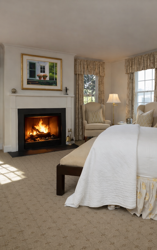 Light-filled bedroom at The Fearrington House Inn with a white bed, upholstered chairs, and a lit fireplace beneath a framed artwork, styled in soft neutral tones.