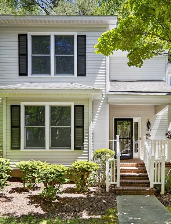 Two-story light gray townhouse with white trim and black shutters, surrounded by mature trees and neatly maintained landscaping. A brick staircase leads to a covered front porch with white railings and a glass-paneled front door, while large windows bring natural light into the home. The shaded front yard features trimmed shrubs, mulch beds, and a concrete walkway leading from the sidewalk to the entrance.