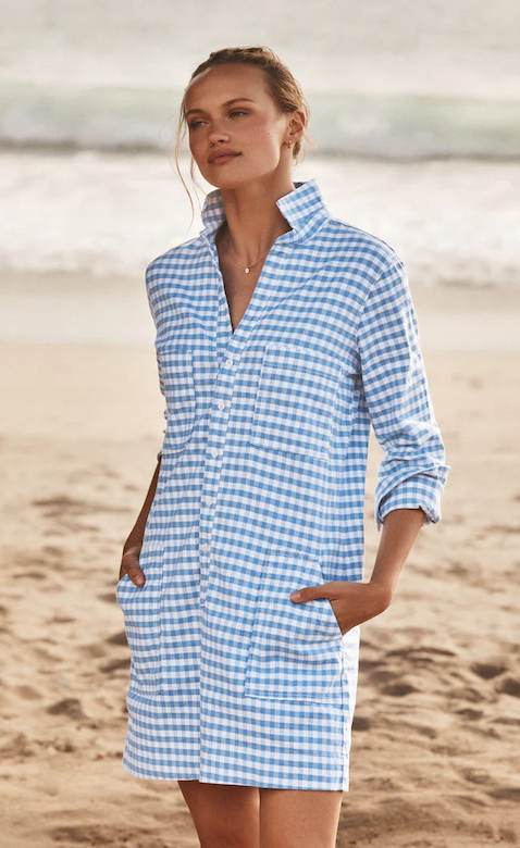 Woman standing on a sandy beach wearing a relaxed, blue-and-white gingham shirt dress with a popped collar and front pockets. Hands in pockets, with soft waves and shoreline in the background, creating a breezy, coastal feel.