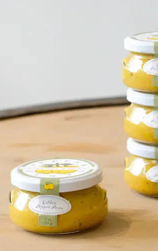 Small glass jars of golden pepper pesto with white lids stacked on a wooden surface.