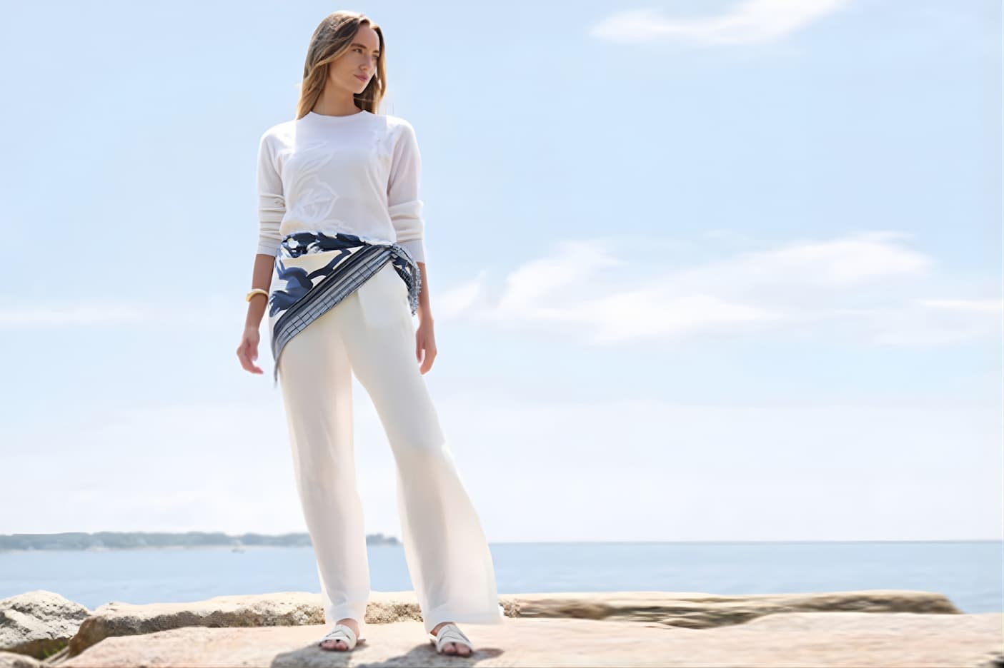Woman standing on seaside rocks wearing a white sweater and wide-leg trousers with a patterned scarf tied at the waist, ocean in the background.