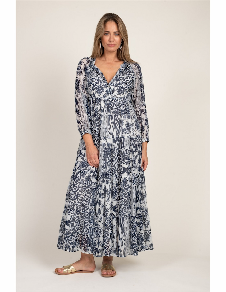 Woman wearing a long, flowy blue-and-white patterned dress with three-quarter sleeves and a tiered skirt, paired with flat sandals against a neutral background.