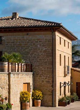 A sunlit stone building with a tiled roof and simple shutters stands along a quiet street, accented by potted plants and warm-toned stucco details.