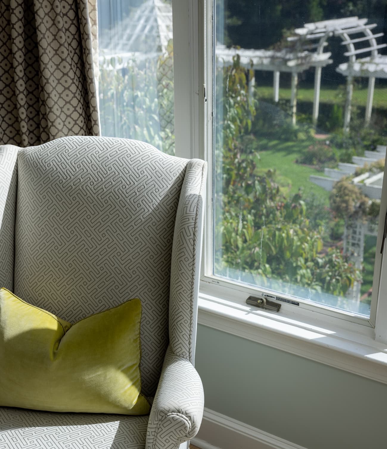 Sunlit wingback chair with a soft green accent pillow beside a window overlooking Fearrington's gardens, pergola, and landscaped grounds.