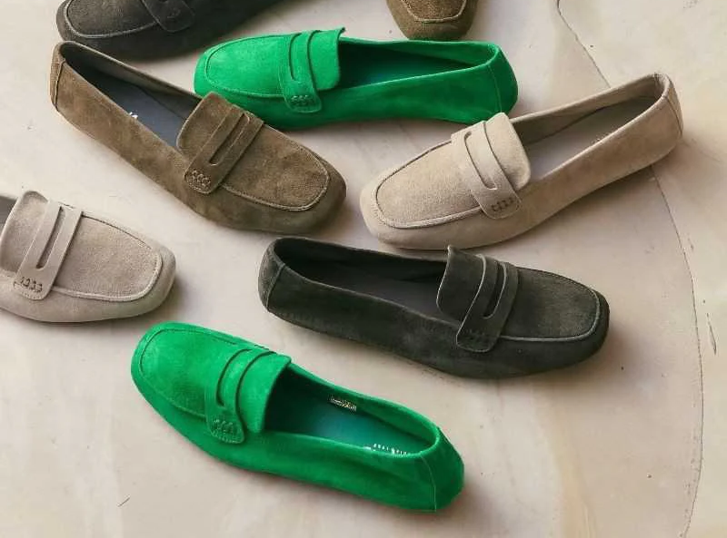 Assorted suede loafers in green, taupe, beige, and charcoal arranged casually on a pale surface, highlighting soft texture and classic penny-loafer styling.