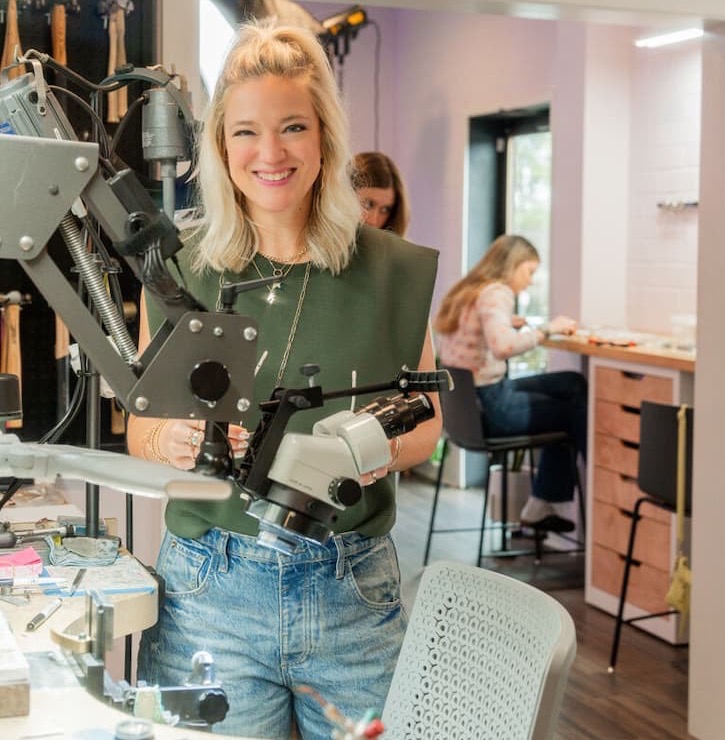A smiling jeweler stands at a workbench holding specialized equipment, with tools and machinery in the foreground and others working at benches in a bright studio behind her.