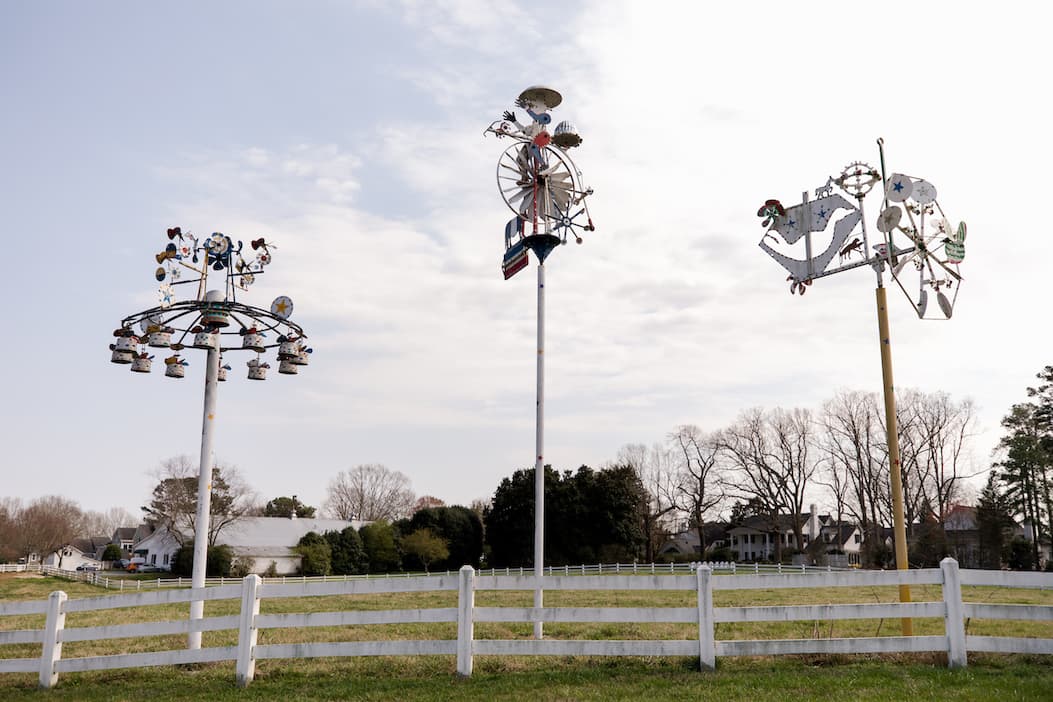 Three large metal whirligig sculptures on tall poles in an open field with houses and trees in the background.
