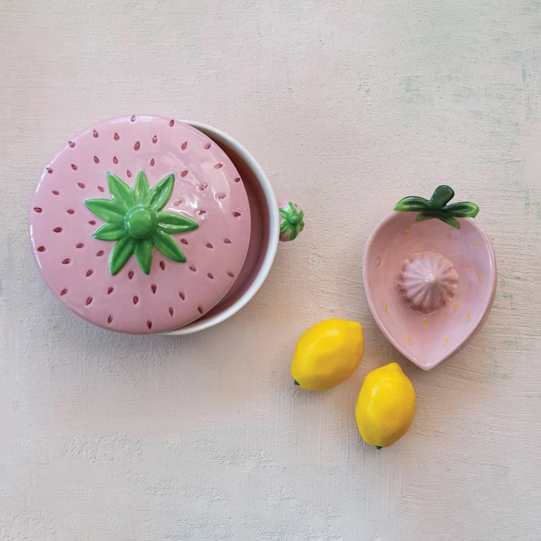 Overhead view of a whimsical pink strawberry-themed ceramic set on a softly textured surface. A strawberry-shaped covered container sits beside a matching strawberry-shaped citrus juicer, accented with glossy green leaf details and accompanied by two bright yellow lemons for a cheerful, playful display.