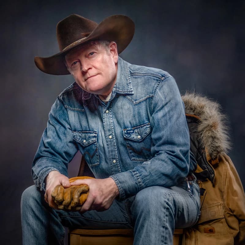 Man in a wide-brimmed cowboy hat and denim shirt sits leaning forward, hands clasped in work gloves. A rugged jacket is draped behind him, and the studio backdrop creates a classic, outdoorsman portrait feel.