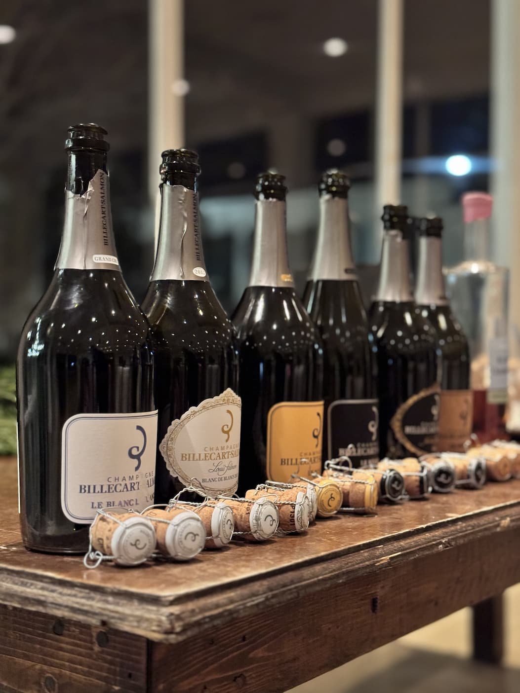 A lineup of Champagne bottles and corks is displayed on a rustic wooden table during a tasting event. Warm indoor lighting and softly blurred windows in the background create an elegant atmosphere, highlighting a selection of sparkling wines arranged for comparison and enjoyment.
