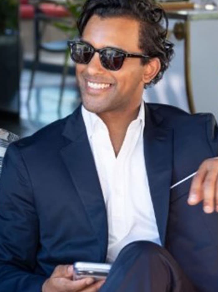 Man in a navy suit and white shirt, wearing sunglasses, smiling while seated and holding a phone outdoors.