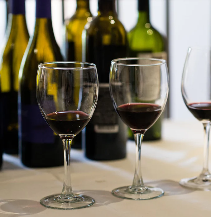 Two wine glasses partially filled with red wine on a table, with wine bottles blurred in the background.