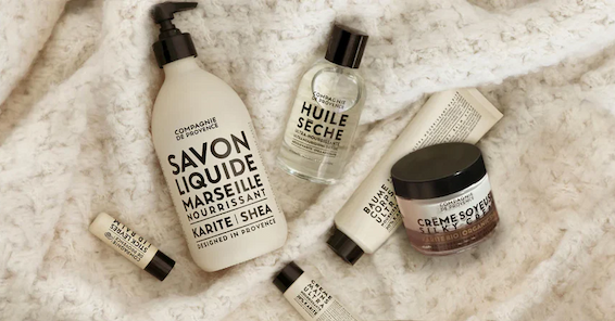 Flat lay of French-inspired bath and body products arranged on a soft white textured blanket, featuring liquid Marseille soap, dry oil, hand creams, and a jar of body cream in clean, neutral packaging.