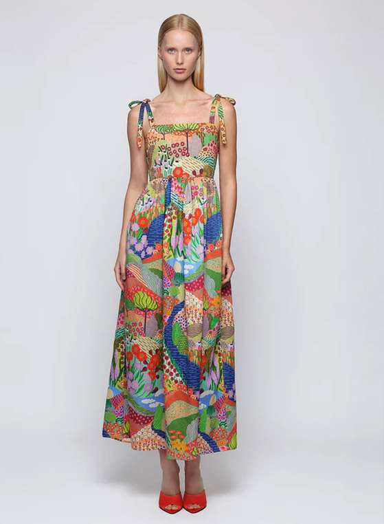 Woman wearing a colorful printed midi dress with tie shoulder straps and a square neckline, featuring a vibrant botanical landscape pattern in shades of coral, green, blue, and pink, paired with red heels against a neutral studio backdrop.