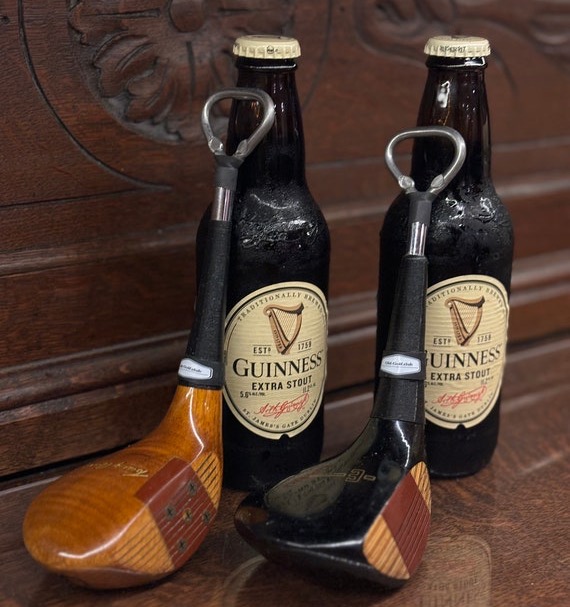 Two handcrafted golf club bottle openers lean against bottles of stout, highlighting their polished wooden club heads, leather-look grips, and metal opening loops. The vintage-inspired bar accessories combine classic golf style with everyday function.