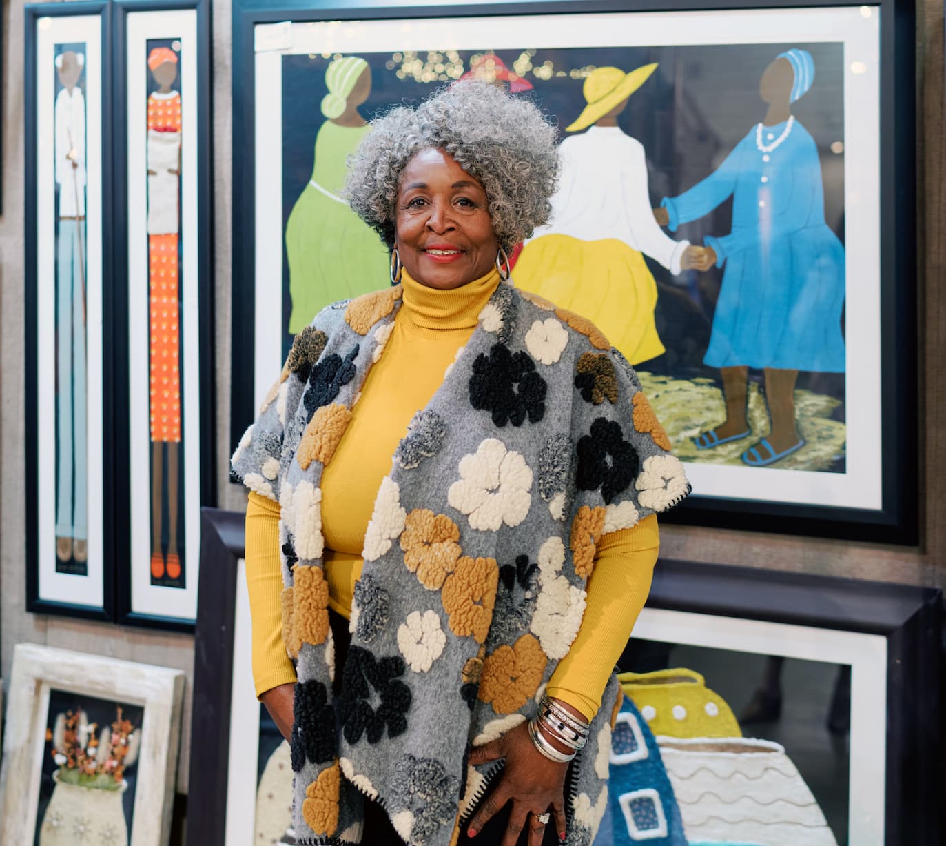 An older woman with curly gray hair smiles while standing in front of framed folk art paintings. She wears a mustard yellow turtleneck and a patterned vest with raised floral textures in neutral tones, accessorized with bracelets, in a gallery or art fair setting.