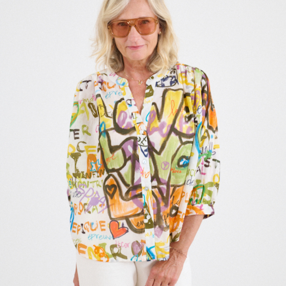 Woman wearing amber sunglasses and a white blouse printed with colorful abstract graffiti-style patterns stands against a light background.