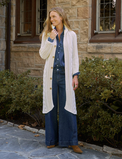 Woman wearing a long cream cable-knit cardigan over a denim shirt and wide-leg jeans, standing outside a stone building. Fearrington Village.