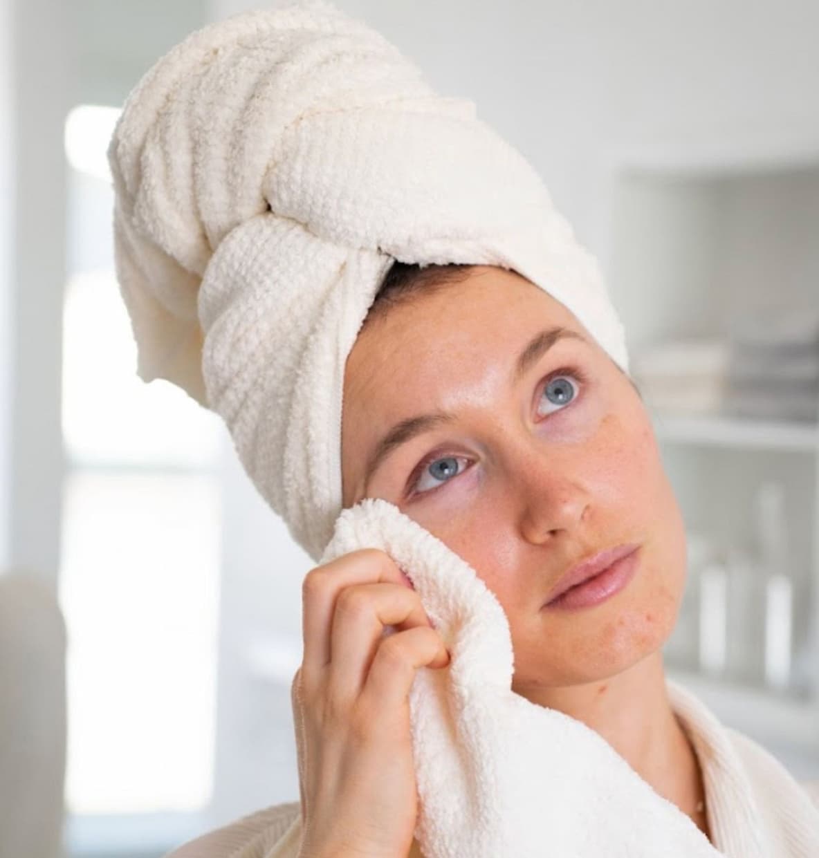 Woman with a towel wrapped around her hair gently patting her face with a soft towel, standing in a bright, minimal bathroom setting.