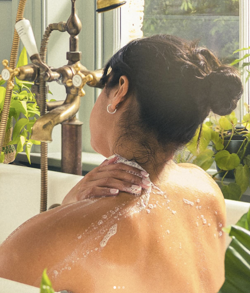 Woman seen from behind bathing in a tub, gently washing her shoulder with soap, with warm light, vintage brass fixtures, and leafy plants by a window creating a calm, spa-like setting.
